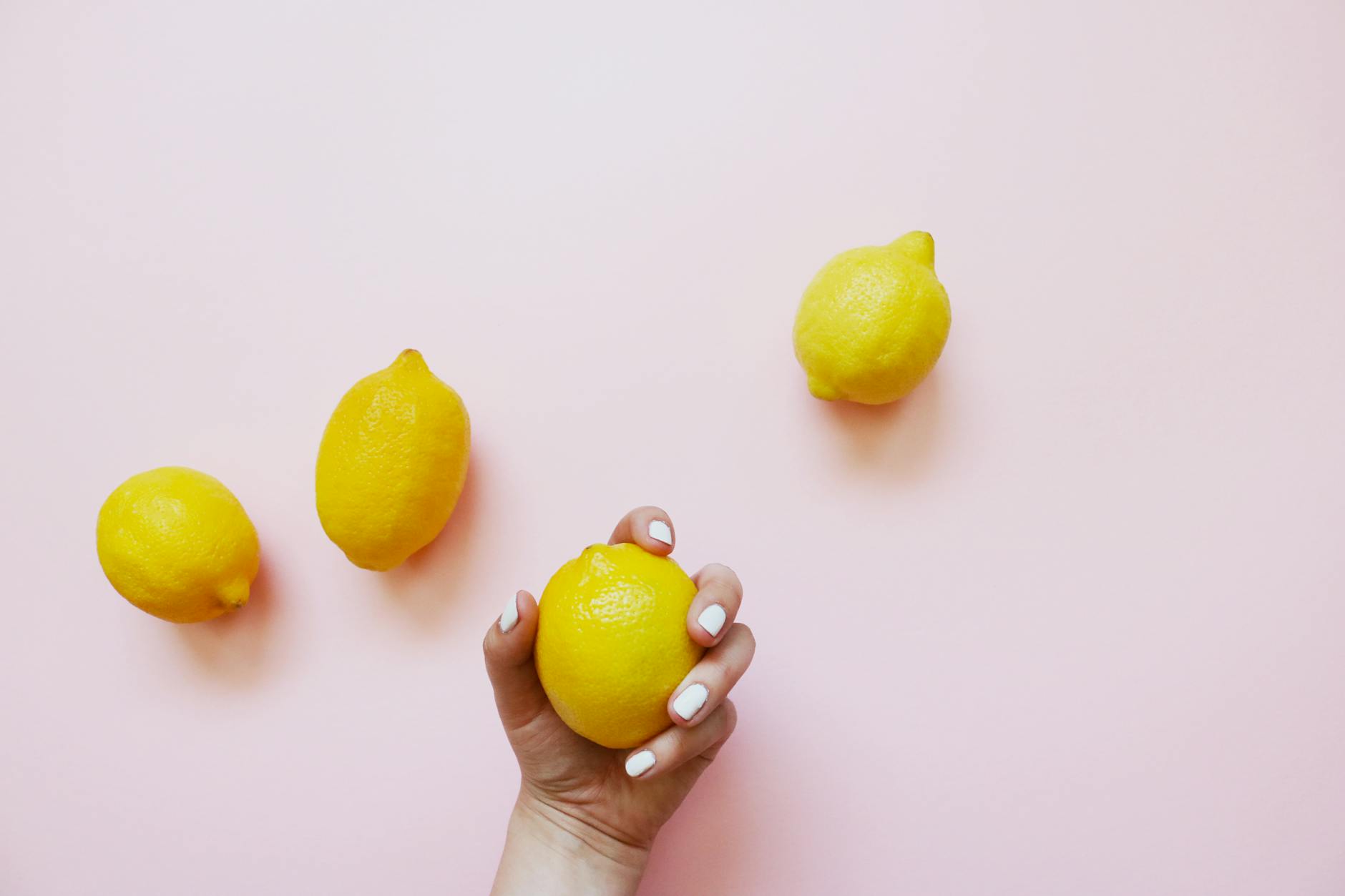 Hand with white nails holding a fresh lemon on a soft pink background with additional lemons nearby