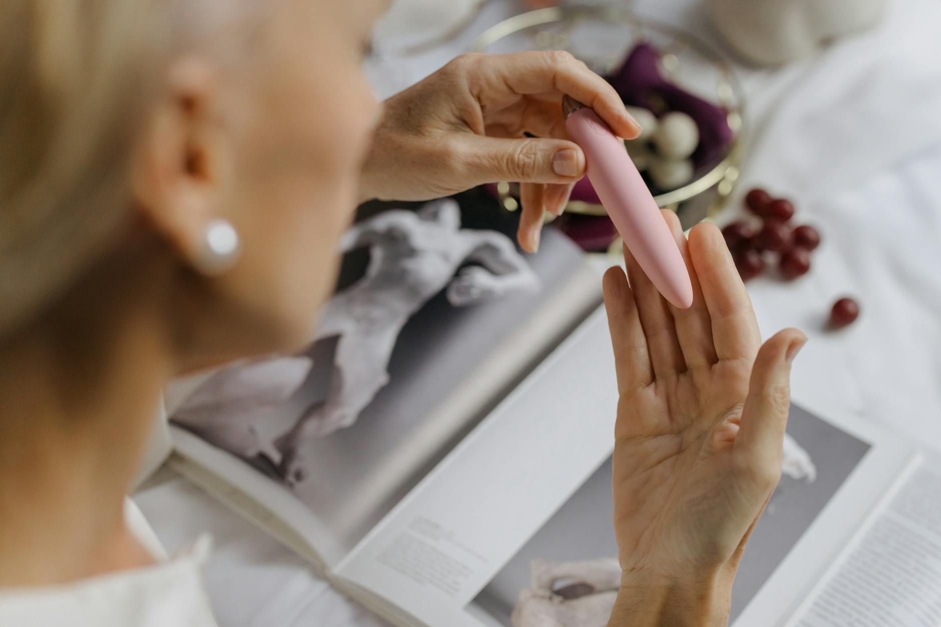 Close-up of a woman's hand holding a pink vibrator, with art books in the soft background, symbolizing personal empowerment and healing